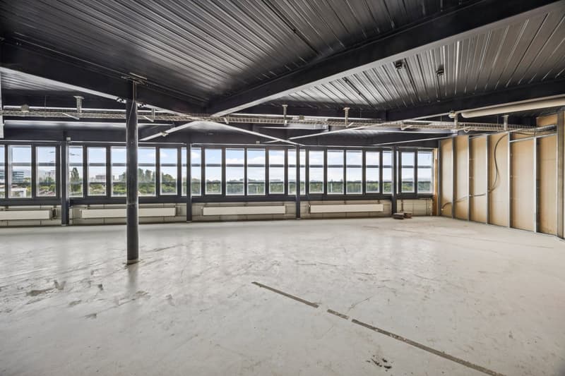 Large open space with high ceilings, concrete floor, and large windows offering a panoramic view of the surrounding area. The space appears to be a commercial or industrial building, possibly an office or warehouse.