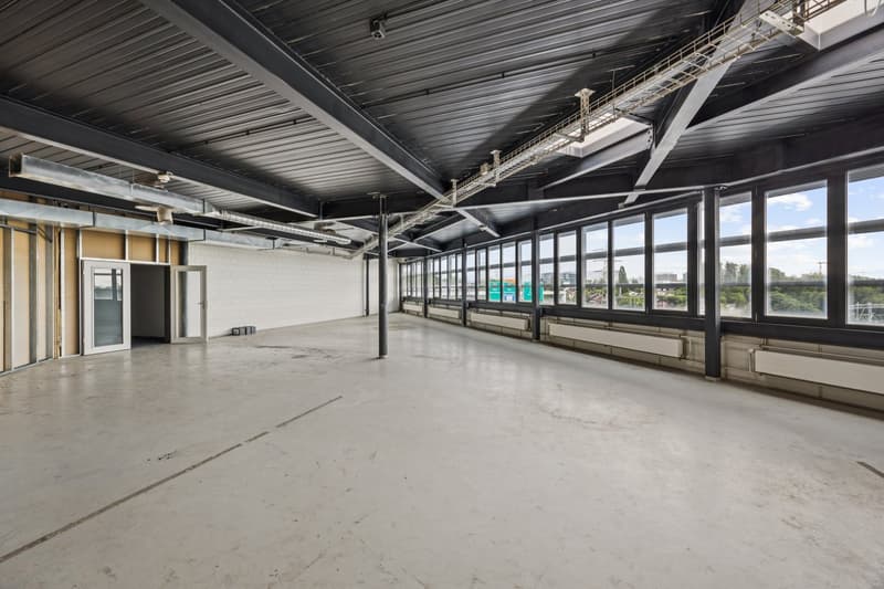 Large open commercial space with high ceilings, exposed metal beams, and large windows providing a view of the surrounding area. The space appears to be unfinished or under renovation, with a concrete floor and minimal furnishings.