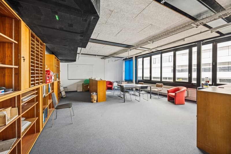 Large open office space with wooden shelving units, tables, chairs, and a projector screen. The room has large windows providing natural light and a modern, industrial-style ceiling.