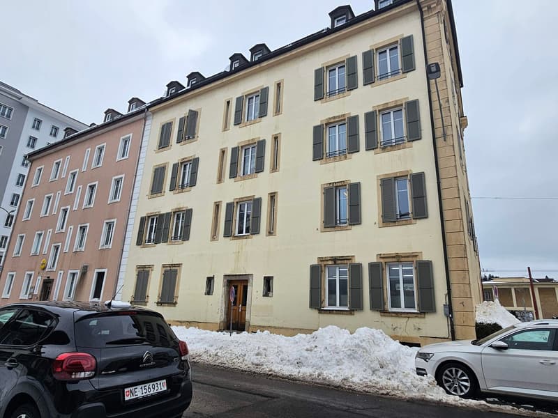 An apartment building with yellow walls, numerous windows, a brown entrance door, two parked cars on the side, and snow cover