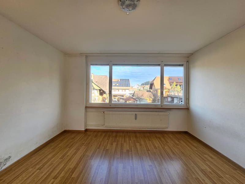 An empty room with white walls, wooden floor, large window, and a radiator beneath the window.