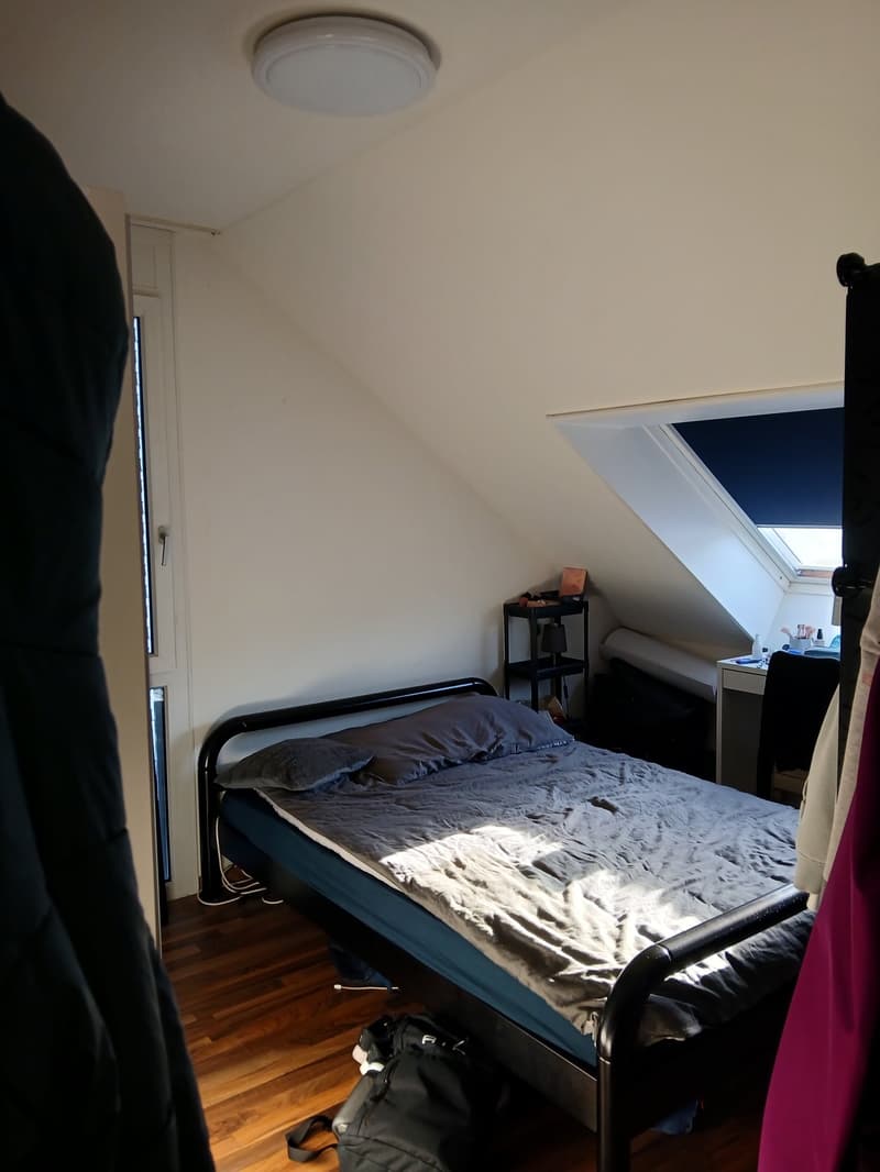 small single bed, bed sheets and pillows, table with lamp, clothes hanging from rack