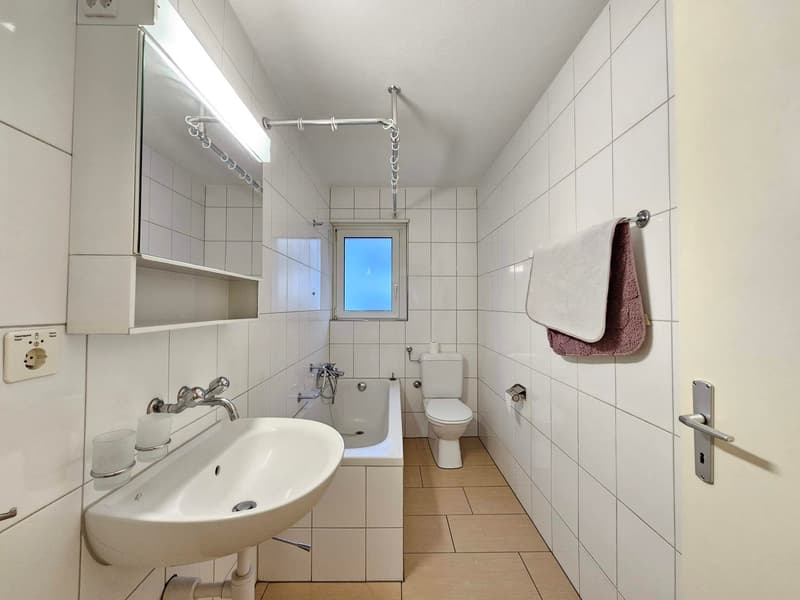 Bathroom with white tiled walls and floor, bathtub, toilet, sink, mirror, towels on towel rack, power outlet, door.