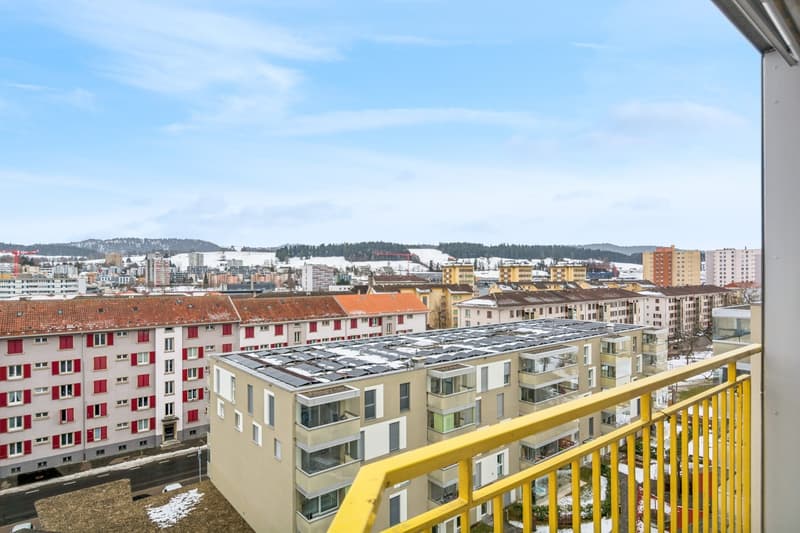 Multiple residential buildings, a balcony with a view, yellow railings, red shutters on windows, snow-covered rooftops, distant mountains.