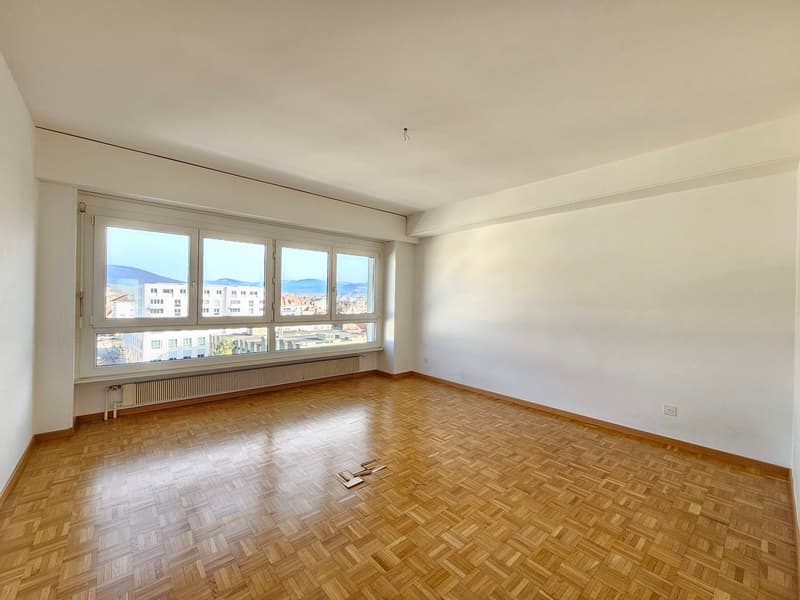 Empty room with wooden parquet flooring, large window with a city view, white walls, and a radiator below the window.