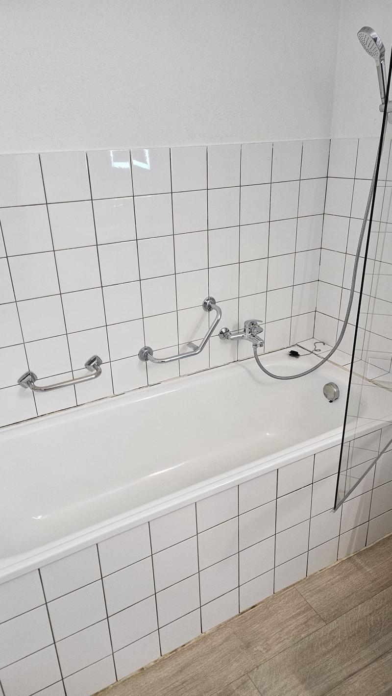 white bathtub with shower, tiled walls, white tiles, wooden floor, metal handrails