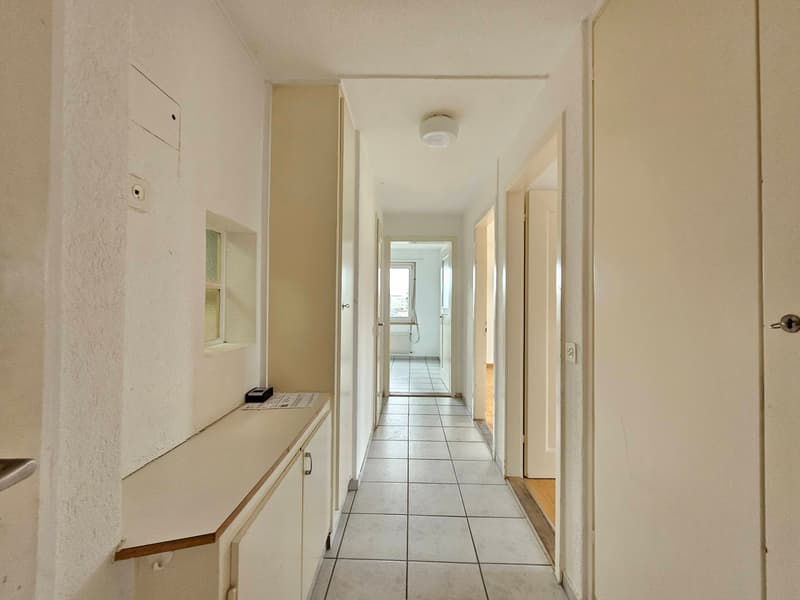Empty hallway with white tiles, white walls, and doors on both sides. A small counter with cabinets is on the left side.