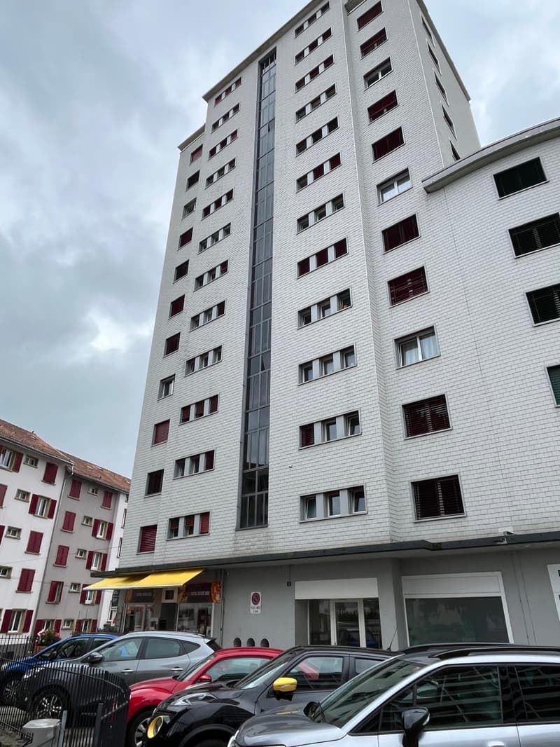 A tall white building with many windows, red shutters, and a yellow awning. Several cars are parked in front of the building.