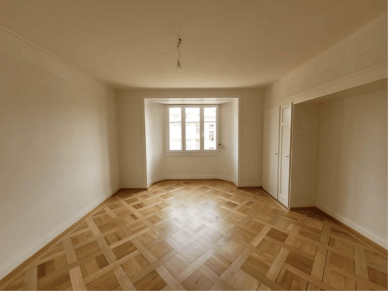 Empty room with wooden parquet flooring, white walls, and a window. No furniture or equipment.