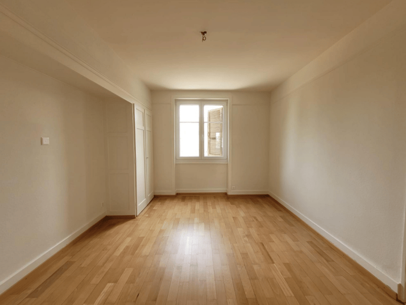 Spacious room with white walls, hardwood flooring, and a large window providing natural light. The room appears to be empty, ready for furnishing and decorating.