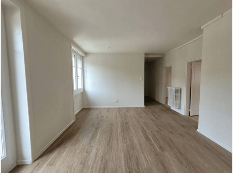 An empty room with white walls, wooden floors, and a window. There are no visible objects or furniture.
