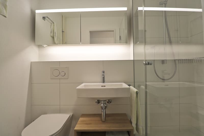 Modern bathroom with white tiles, modern white sink, under sink storage, walk-in shower with glass enclosure, modern faucets and shower head, mirror with integrated lights, towel rack, white toilet.
