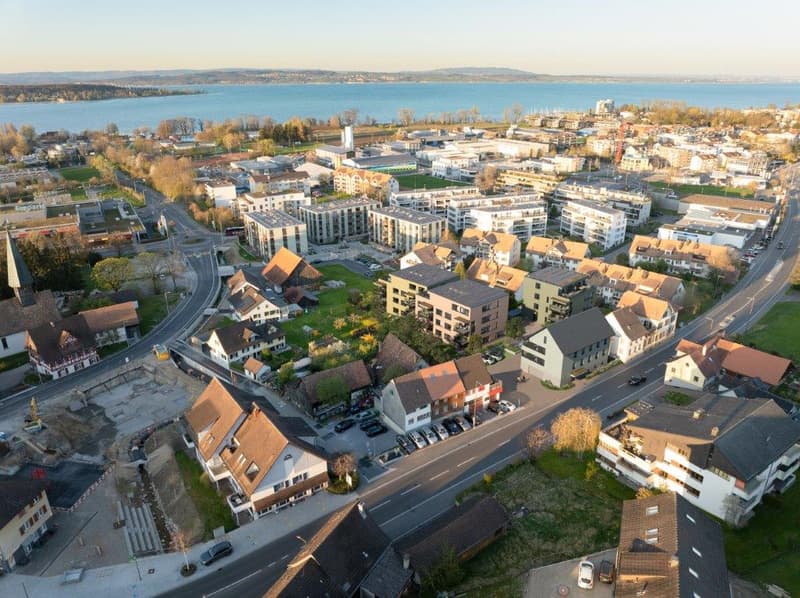 Aerial view of a city with a mix of residential and commercial buildings, surrounded by a large body of water and hills in the background. The city appears to have a mix of modern and older architecture, with a variety of building heights and styles. Ther