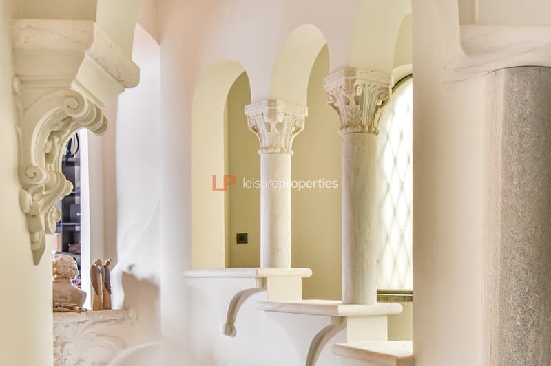 Staircase leading up to an upper floor. Features a decorative cornice, arched passageways, and ornate columns. The walls are painted in light colors, and there are small sculptures on a shelf to the left.