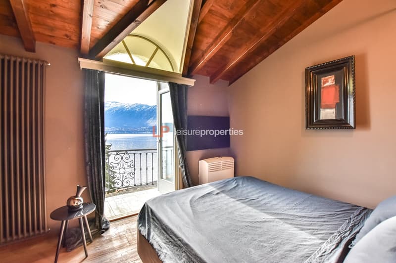 bedroom with balcony and lake view, wooden floor, white wall, bed with grey sheet and pillows, small side table, radiator, picture frame on the wall