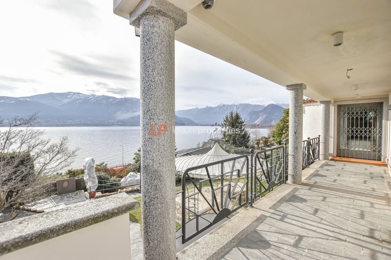 The entrance porch has two pillars with a black railing, a door, and a view of a body of water, mountains, and a house with a gazebo in the distance.