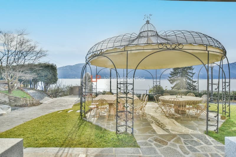 a metal gazebo with a stone floor in the garden with a large green lawn overlooking a beautiful lake and mountains