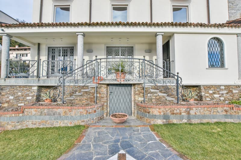 white house, stone facade, balcony with wrought iron railing, central entrance with double door, stone steps leading up to entrance, two potted plants on front steps, decorative stonework