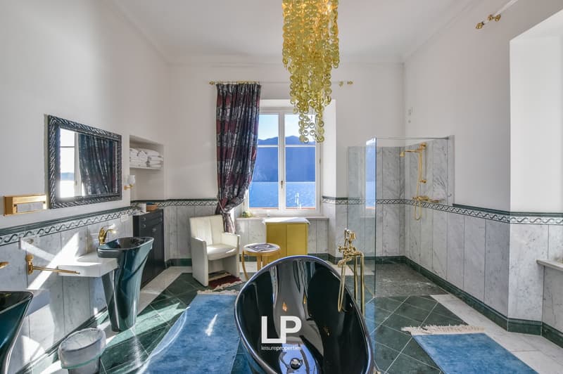 Modern bathroom, marble tiles, white walls, glass shower, curved tub, gold faucets, large mirror, blue rug, white chair, small table, chandelier, view of the sea