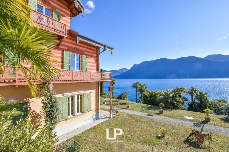 Lake Como Villa, red facade, green shutters, two floors, balcony with decorative railing, green grass, patio with potted plants, and lake view.
