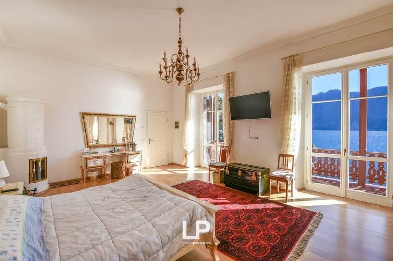 bedroom with bed, wooden floor, mirror, chandelier, fireplace, desk, windows, tv, balcony, red rug, armchairs