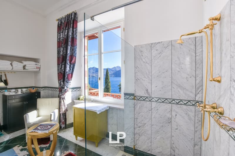 White marbled tiles, glass shower enclosure, golden shower head, white furniture, large window, lake view