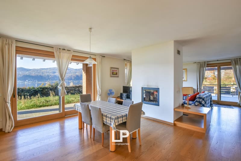 Spacious open-plan living and dining area with large windows offering a scenic view of the surrounding landscape. The room features hardwood flooring, a dining table with chairs, and a cozy seating area with a fireplace. The space is bright and airy, crea