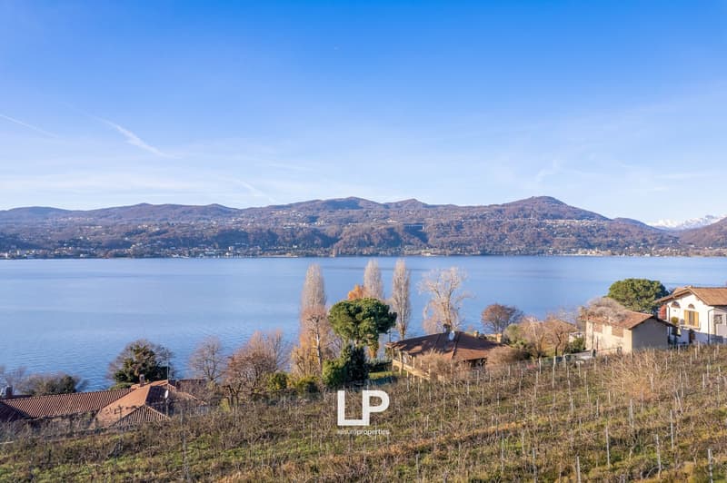 The image shows a scenic view of a lake surrounded by mountains and hills. There are several buildings and houses visible on the hillsides, along with trees and vineyards. The sky is clear and blue, creating a picturesque landscape.