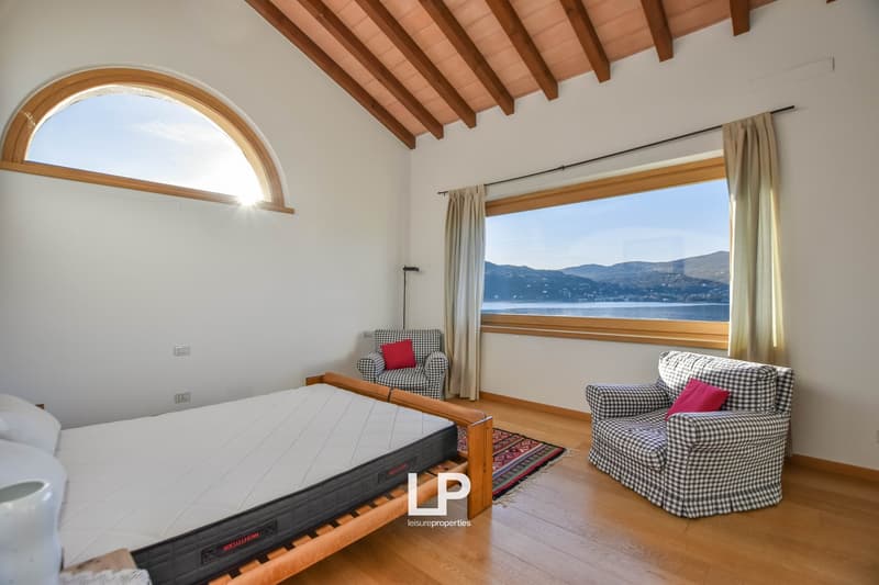 Spacious bedroom with wooden beamed ceiling, large arched window overlooking a scenic lake and mountains, wooden furniture including a bed and armchairs, and a rug on the hardwood floor.