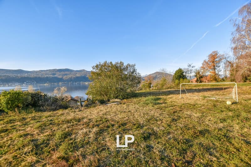 The image shows a scenic outdoor landscape with a lake or body of water in the background, surrounded by hills and mountains. There are trees and vegetation in the foreground, and a soccer goal post can be seen on the grassy field. The sky is clear and bl