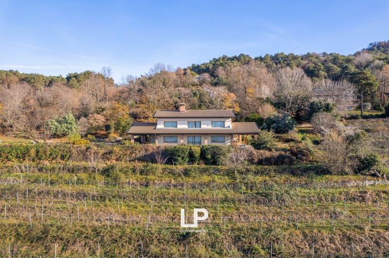2-story house with a tiled roof, surrounded by trees and vegetation on a hillside. The house has several balconies or terraces and offers a scenic view of the surrounding landscape.