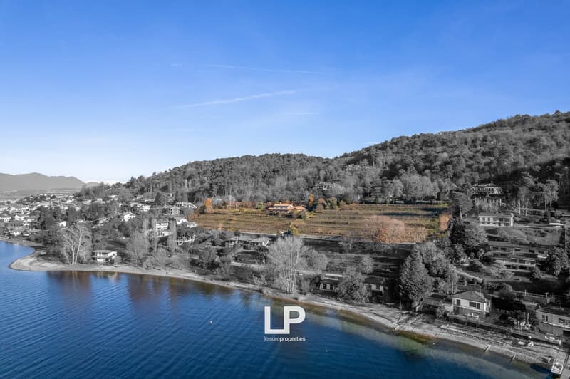 The image shows a scenic landscape with a lake or river in the foreground, surrounded by hills and mountains covered in trees. There are several buildings and houses visible in the distance, nestled among the natural surroundings. The sky is a clear blue,