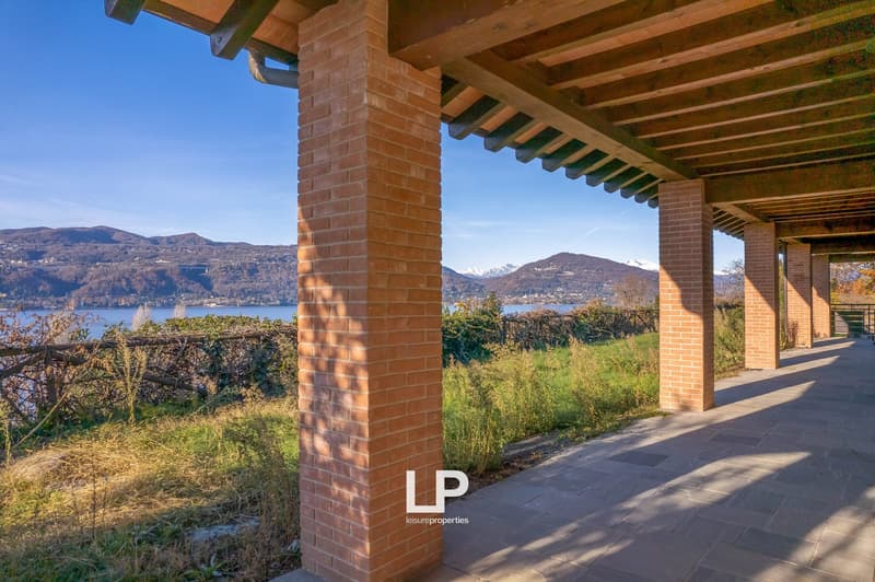 Two-story brick house with wooden beams, large windows, and a covered patio or balcony overlooking a scenic lake and mountain landscape