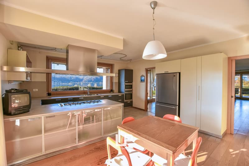 Spacious open-plan kitchen and dining area with large windows offering a scenic view. The kitchen features modern appliances, including a refrigerator and gas stove, as well as a central island with a wooden countertop. The dining area has a wooden table