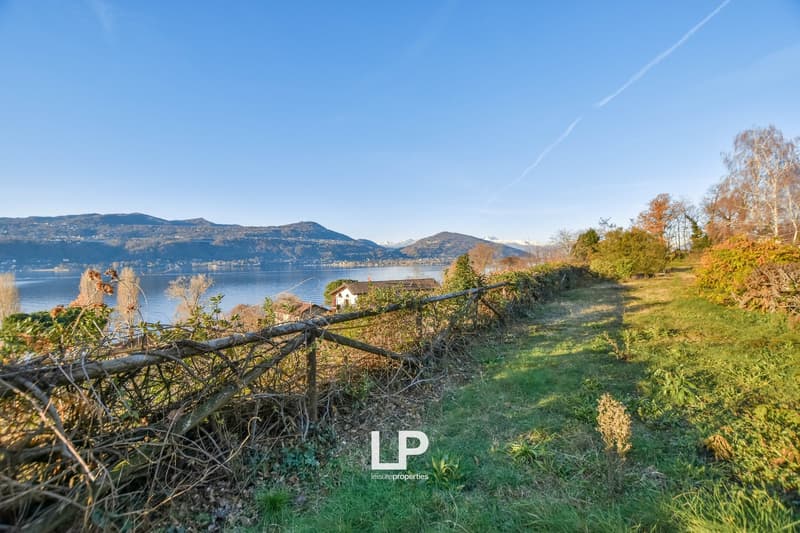 The image shows a scenic landscape with a lake, mountains, and a small house in the distance. The foreground features a grassy path with a wooden fence and autumn-colored trees. The sky is clear and blue, with a few contrails visible.