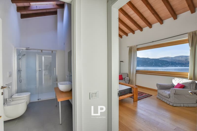 This image shows a bedroom or living space with a stunning view of the lake and mountains through the large windows. The room has a wooden ceiling with exposed beams, hardwood floors, and a cozy seating area with a sofa. There is also a bathroom with a gl