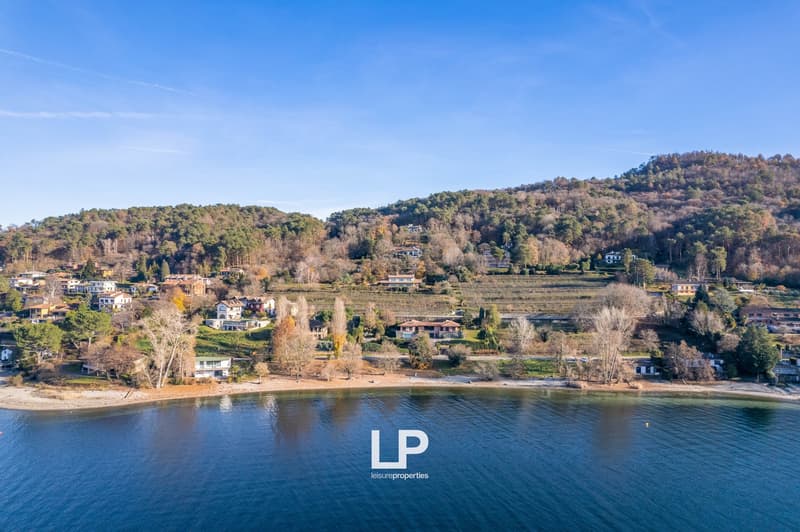 The image shows a scenic landscape with a lakefront view. The landscape features rolling hills covered in autumn foliage, with a mix of residential and natural areas. There are several buildings and houses visible along the shoreline. The lake itself is a