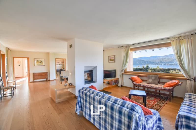 Spacious living room with hardwood floors, large windows offering a scenic view of the lake and mountains, a fireplace, a plaid sofa, and a wooden entertainment unit with a TV.