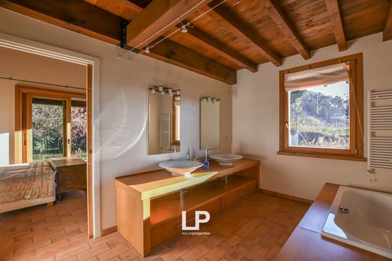 Spacious bathroom with wooden beamed ceiling, double vanity sinks, large window overlooking outdoor area, and access to a balcony or terrace