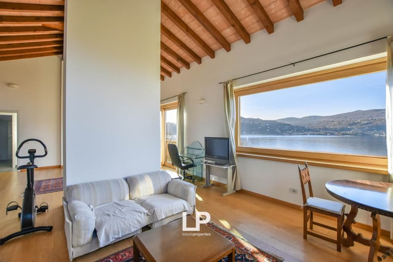 Spacious living room with wooden beamed ceiling, large windows overlooking a lake, sofa, TV, and a small dining area.