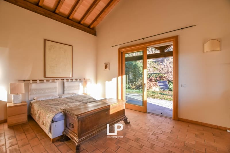 Spacious bedroom with wooden beamed ceiling, large windows overlooking a garden, wooden furniture including a bed and nightstands, and a wooden chest at the foot of the bed.