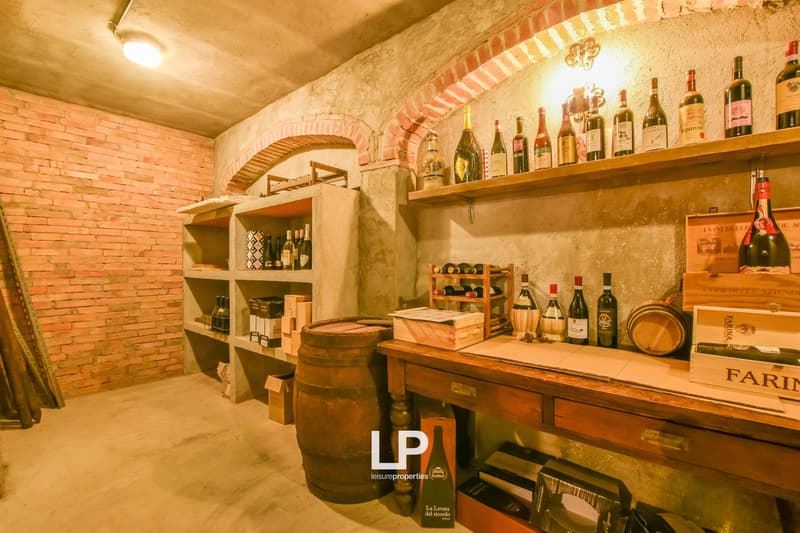 A room with brick walls and shelves full of wine bottles, wooden barrel, wooden table, wooden crates, and lamps on the ceiling.
