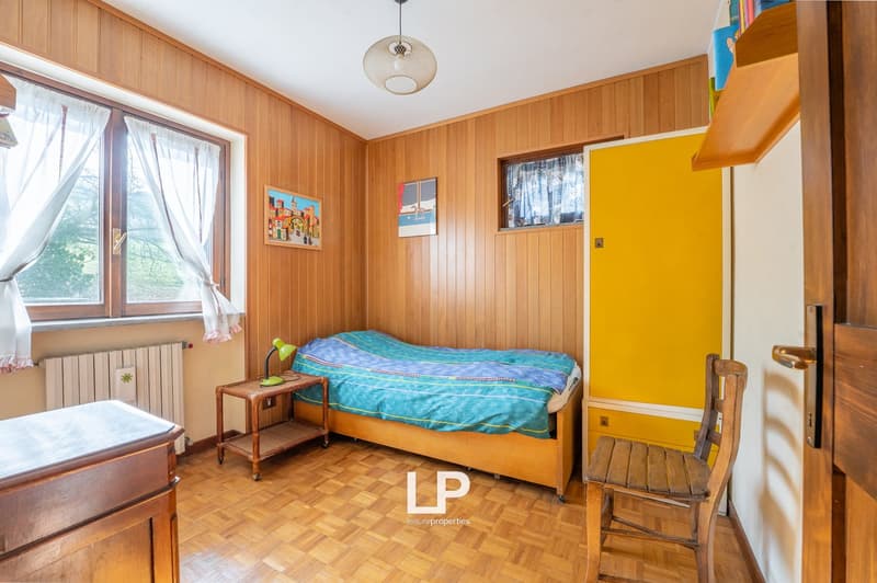small bedroom with bed, nightstand, lamp, wooden chair, wooden wardrobe, wooden wall paneling, radiator, window with curtains