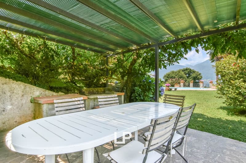 Outdoor dining table with chairs under a pergola, surrounded by lush greenery, a swimming pool, and a distant mountain view.