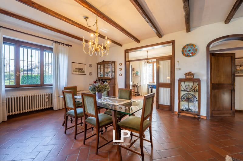 Dining room with wooden table, chairs, chandelier, wooden floor, white curtains, radiator, wooden cabinet, glass cabinet, plate, and picture frame.