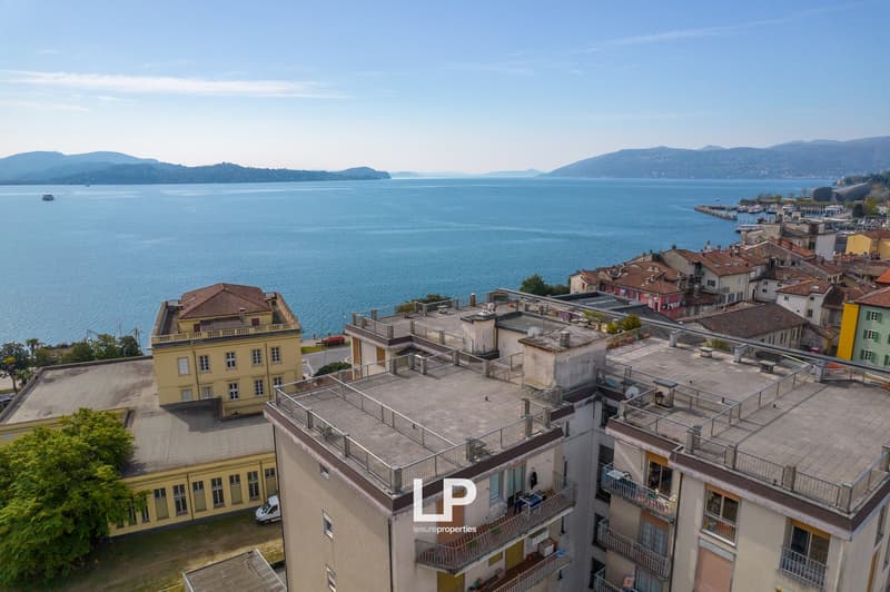 The image shows a scenic view of a coastal city with a large body of water, likely a lake or bay, surrounded by mountains in the background. The foreground features a mix of residential and commercial buildings, some with balconies or terraces visible. Th