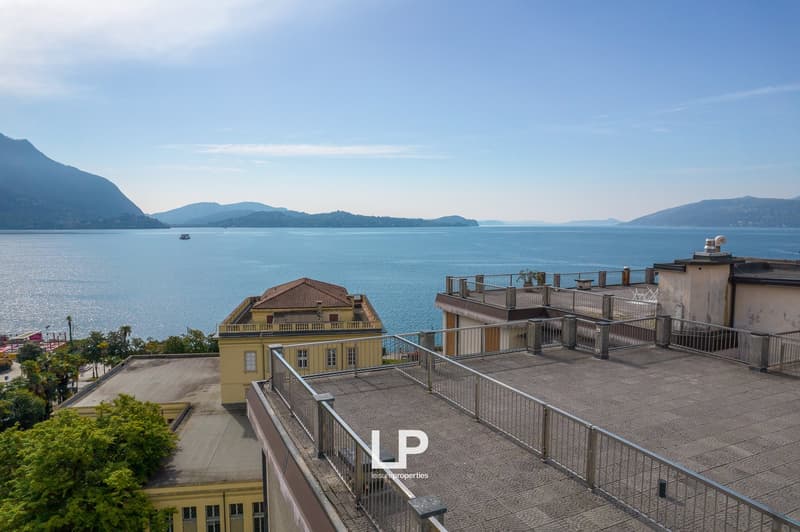 Multi-story yellow building with balconies overlooking a large lake surrounded by mountains. There is a terrace or balcony area on the roof of the building. The image shows a scenic lakeside view with a boat on the water.