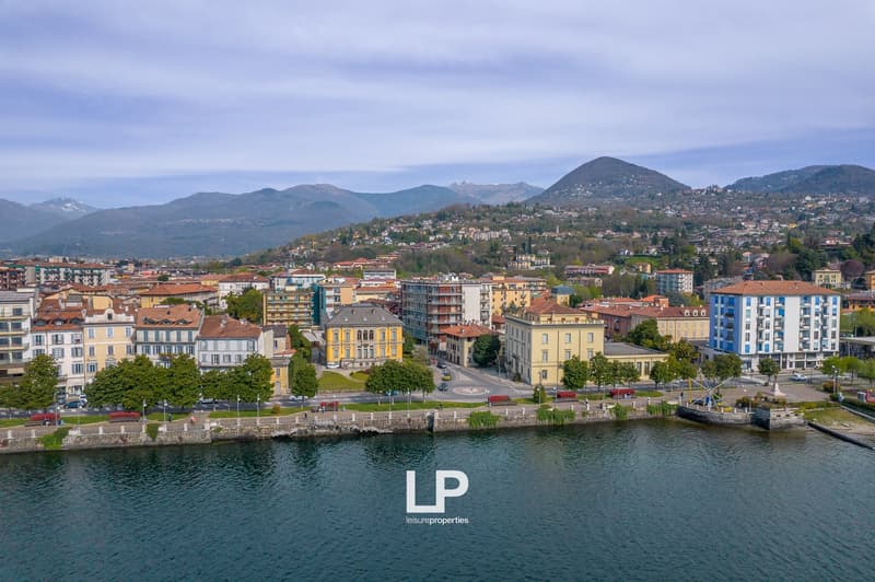 Aerial view of a city located on the shore of a lake or river, surrounded by mountains. The city features a mix of residential and commercial buildings, with a variety of architectural styles. There are trees and greenery visible throughout the cityscape.
