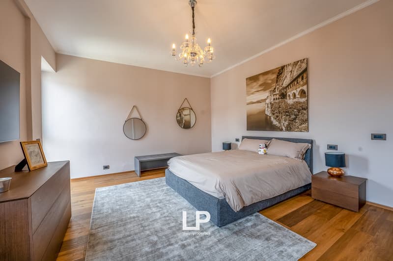 Spacious bedroom with hardwood floors, beige walls, a large chandelier, a gray upholstered bed frame, a wooden dresser, and a framed artwork on the wall depicting a scenic landscape.