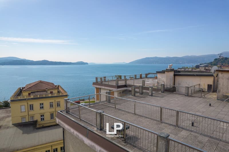 A multi-story building with a balcony and terrace overlooking a large body of water, likely a lake or bay, with mountains in the background. The building has a yellow exterior and appears to be a residential or commercial property.